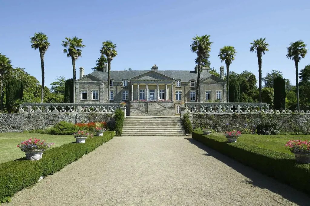 Manoir en pierre au bout d’une allée fleurie bordée de palmiers